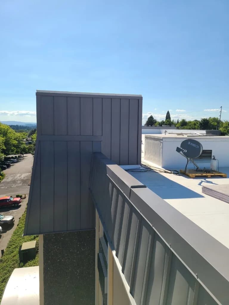High-angle view of Quality Inn in Tigard, OR: charcoal standing-seam parapet and facade wrap meeting a white TPO-style membrane field, with a satellite dish, parking lot, and evergreens beyond