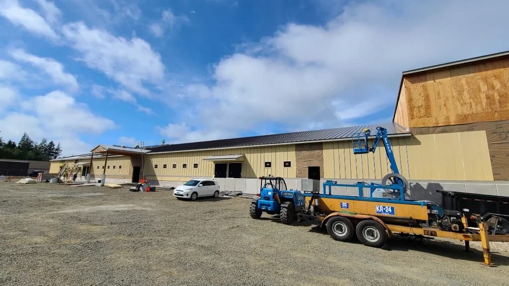 Construction at Roby's Furniture Store in Warrenton, OR: long commercial building with a partial bronze standing seam roof, KR-24 on-site roll former, Genie telehandler, and workers