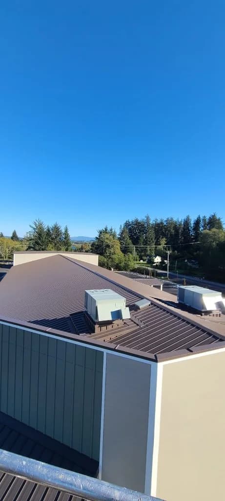 Elevated view of the completed multi-level bronze standing seam roof at Roby's Furniture Store in Warrenton, OR, with HVAC curbs, olive and tan vertical siding, evergreens, and blue sky