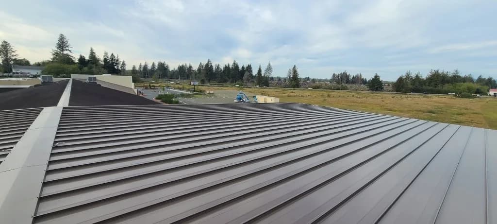 Wide bronze standing seam roof field at Roby's Furniture Store in Warrenton, OR, with a boom lift and Pacific Northwest evergreens beyond