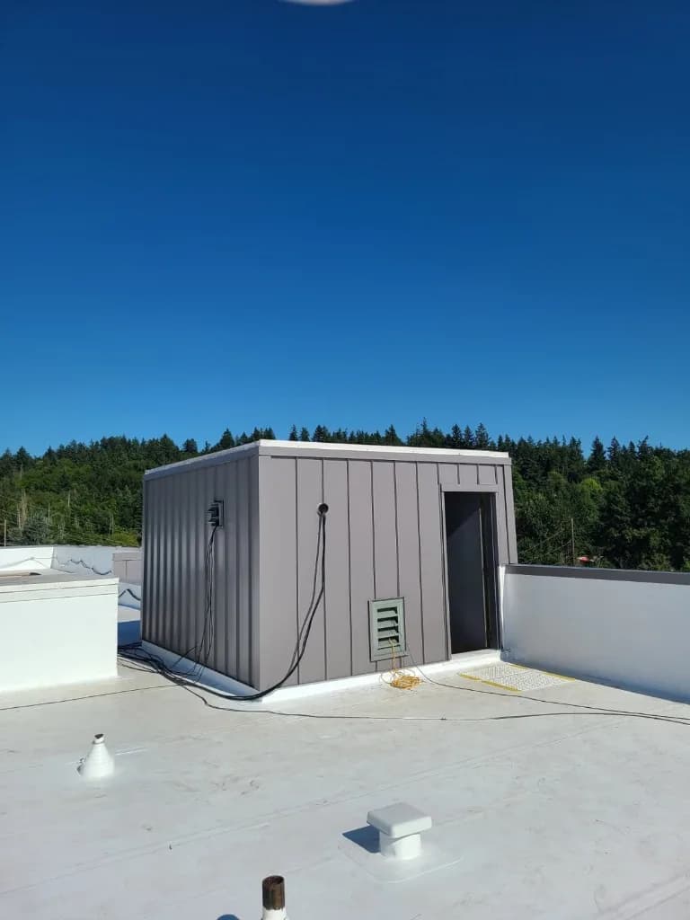 White TPO/PVC-style membrane field with welded seams and roof vents at Quality Inn in Tigard, OR, meeting grey standing-seam penthouse walls under evergreens and blue sky