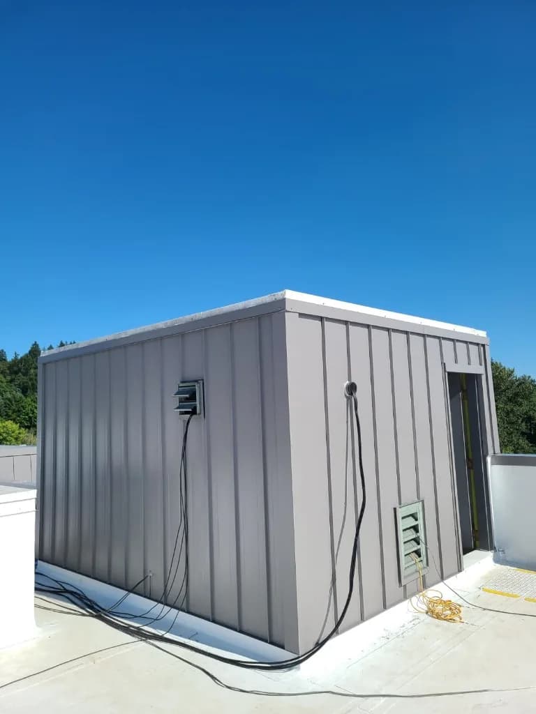 Rooftop equipment penthouse at Quality Inn in Tigard, OR, clad in taupe-grey vertical standing-seam metal with white cap flashing, an open doorway, cables, and a yellow cord on the white membrane